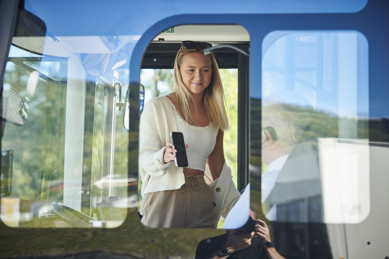 Ung jente på veg inn i bussen. Ho viser billetten som ho har elektronisk på mobilen.
